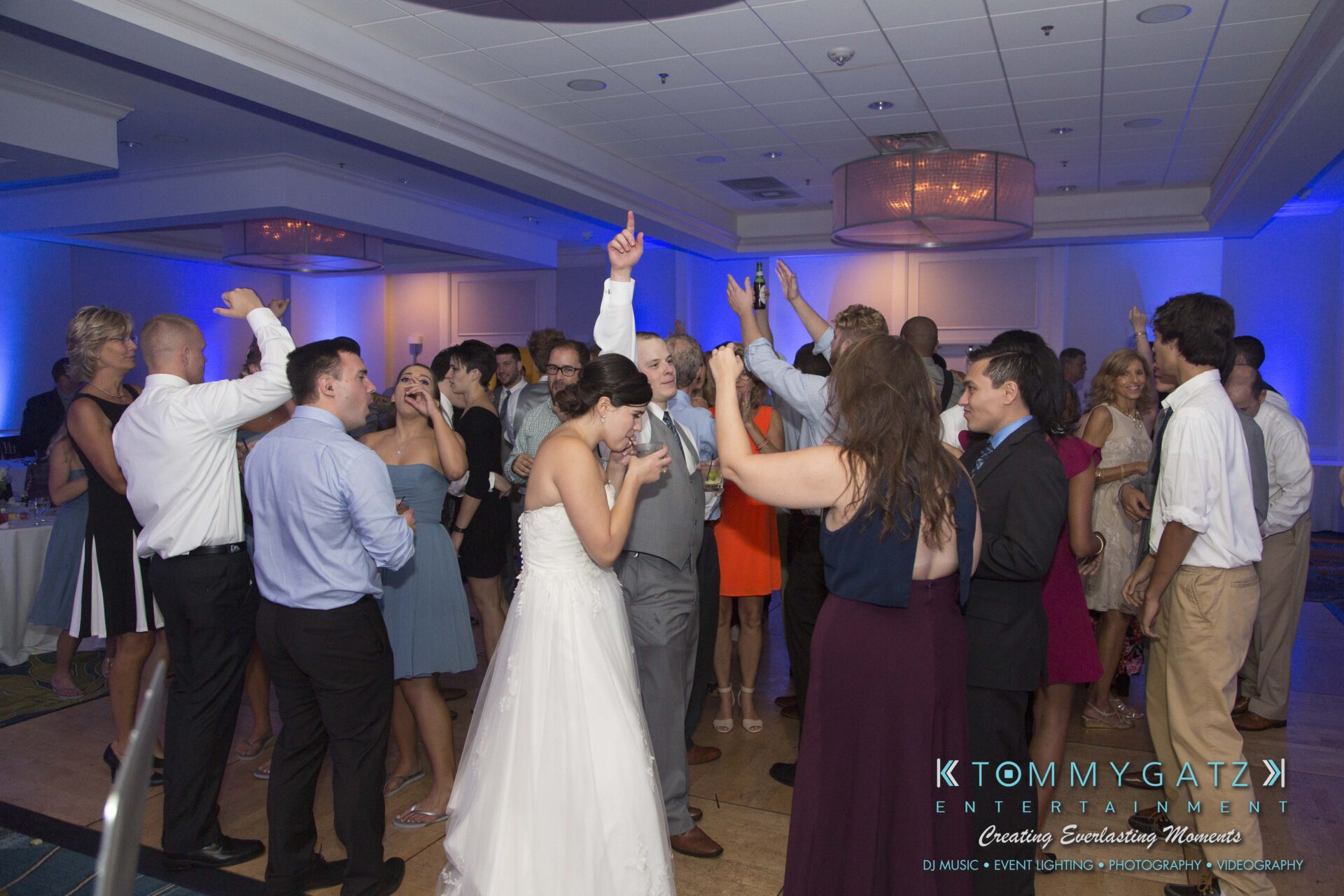 Carly-Patrick-FINAL-Blog-Watermark-51 - TOMMY GATZ ENTERTAINMENT A group of people dancing at a wedding reception in a well-lit banquet hall. A bride in a white dress is at the center of the group.