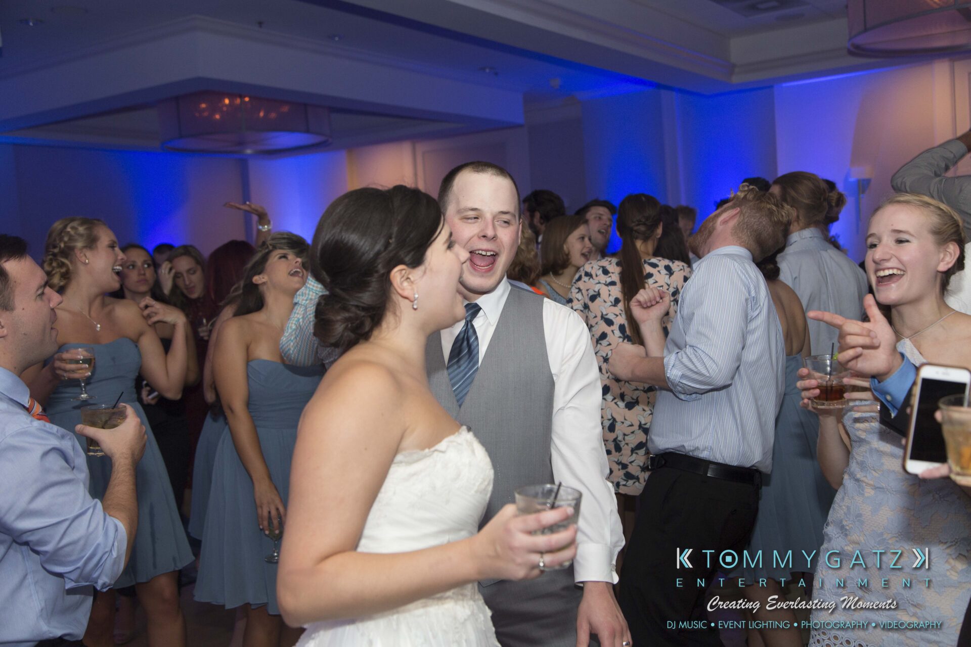 Carly-Patrick-FINAL-Blog-Watermark-52 - TOMMY GATZ ENTERTAINMENT A bride and groom laughing and dancing with guests at a lively wedding reception, with blue lighting and several people holding drinks.
