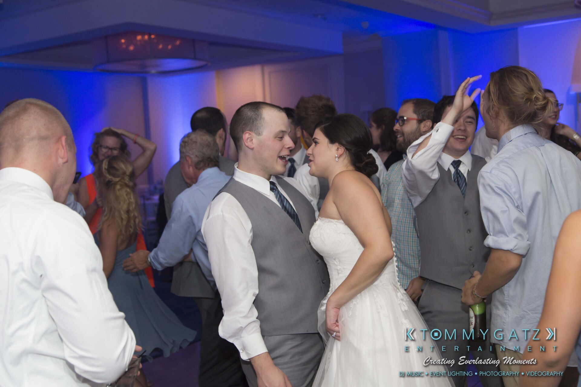 Carly-Patrick-FINAL-Blog-Watermark-54 - TOMMY GATZ ENTERTAINMENT A wedding couple dances closely among guests on a lively dance floor, with blue lighting in the background.