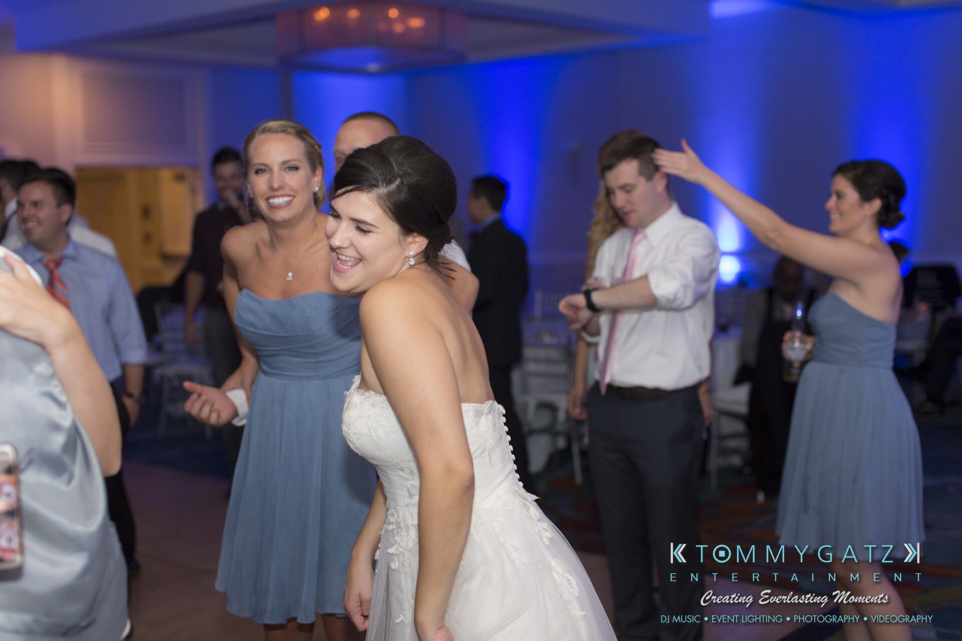 Carly-Patrick-FINAL-Blog-Watermark-56 - TOMMY GATZ ENTERTAINMENT A bride in a white dress smiles and dances in a room with blue lighting, surrounded by guests wearing formal attire.
