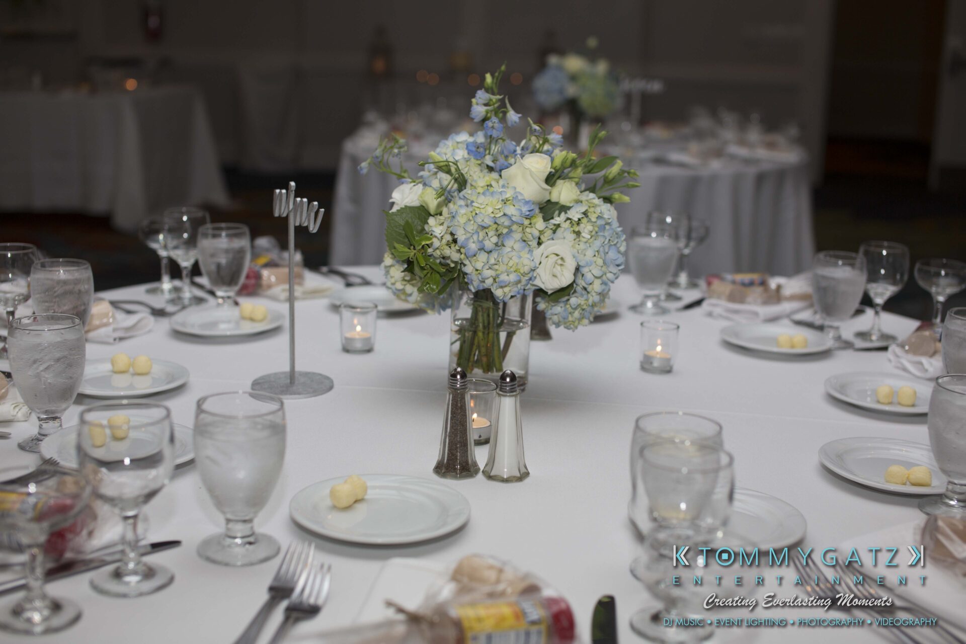 Carly-Patrick-FINAL-Blog-Watermark-7 - TOMMY GATZ ENTERTAINMENT A round table set for a formal event with white tablecloth, floral centerpiece, plates, cutlery, glassware, and small butter servings.