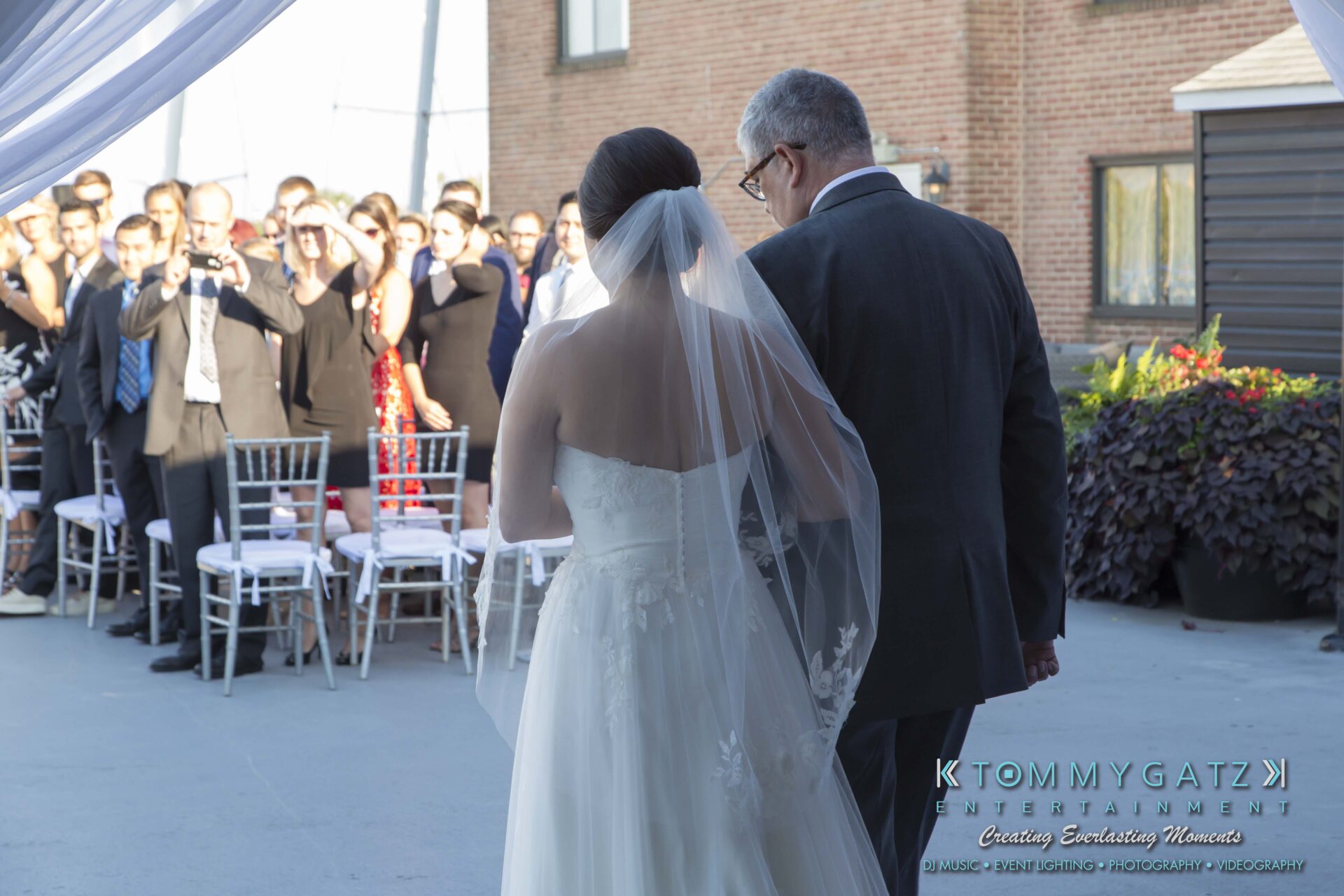 Carly-Patrick-FINAL-Blog-Watermark-8 - TOMMY GATZ ENTERTAINMENT A bride in a white dress walks down the aisle with an older man at an outdoor ceremony. Guests are seated and some are taking photos.
