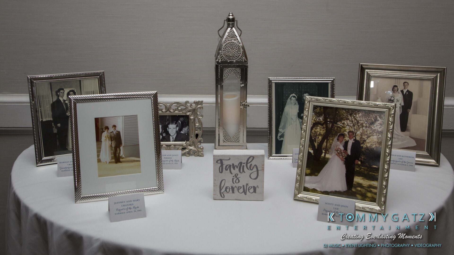 Carly-Patrick-FINAL-Blog-Watermark-9 - TOMMY GATZ ENTERTAINMENT A display of framed wedding photos and a decorative lantern on a round table, with a sign reading "Family is forever.