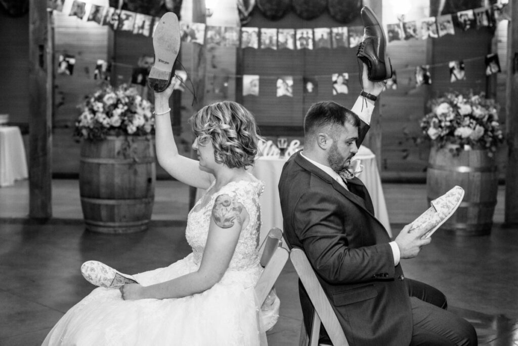 Christine-Jon-Wedding-Blog-100 - TOMMY GATZ ENTERTAINMENT A bride and groom sit back-to-back in chairs, each holding up a shoe. The background features decorative barrels and hanging photos.