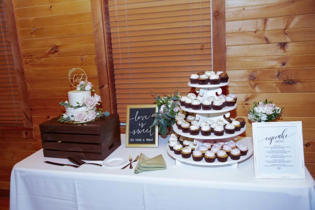 Christine-Jon-Wedding-Blog-101 - TOMMY GATZ ENTERTAINMENT A dessert table with a small decorated cake on a crate, a sign reading "Love is sweet so take a treat," and a multi-tiered cupcake stand next to a framed cupcake menu.