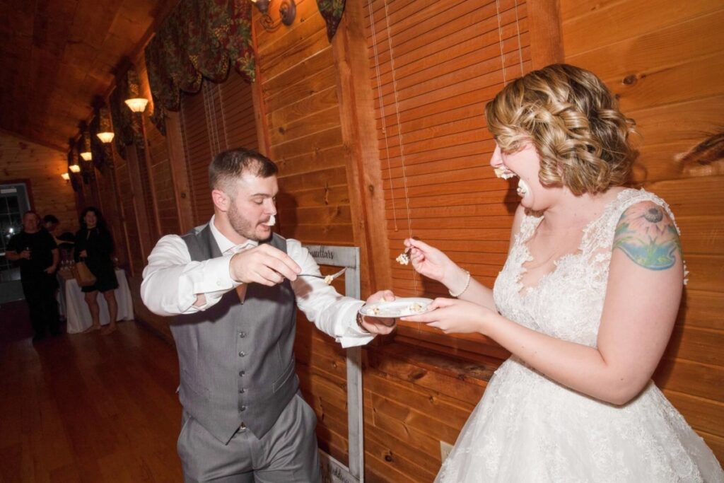 Christine-Jon-Wedding-Blog-102 - TOMMY GATZ ENTERTAINMENT A couple in wedding attire playfully feeds each other cake in a wooden hall.
