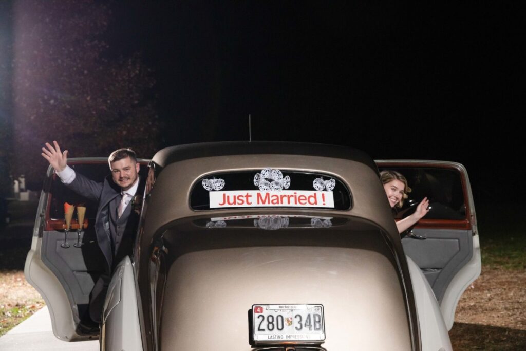 Christine-Jon-Wedding-Blog-104 - TOMMY GATZ ENTERTAINMENT A newlywed couple waves from an open car door, with a "Just Married!" sign on the back of the vintage car at night.