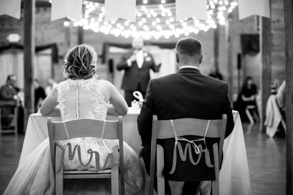 Christine-Jon-Wedding-Blog-95 - TOMMY GATZ ENTERTAINMENT A bride and groom sit at a table labeled "Mr." and "Mrs." facing a speaker. String lights hang above in a decorated venue.