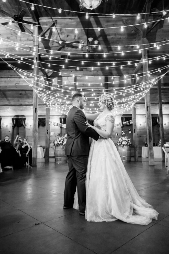 Christine-Jon-Wedding-Blog-96 - TOMMY GATZ ENTERTAINMENT A couple dances in a warmly lit venue with string lights overhead. Guests are seated in the background.
