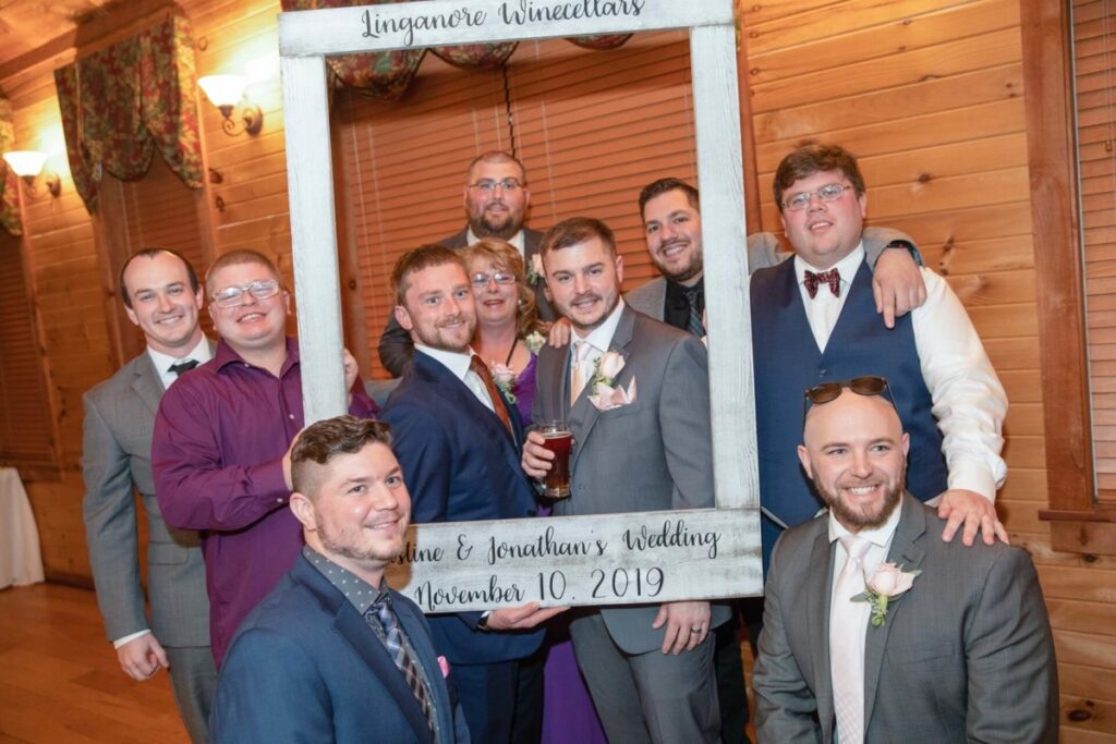 Christine-Jon-Wedding-Blog-98 - TOMMY GATZ ENTERTAINMENT A group of people posing with a large frame at a wedding. The frame reads "Linganore Winecellars" and "Anne & Jonathan's Wedding, November 10, 2019." They are smiling in a wood-paneled room.