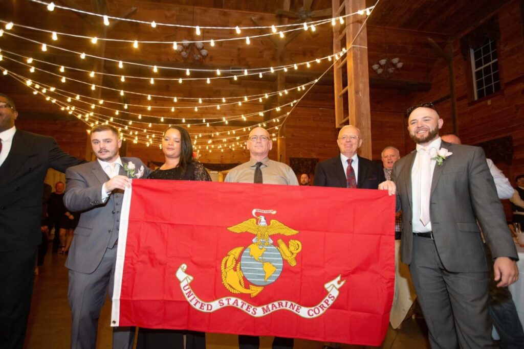 Christine-Jon-Wedding-Blog-99 - TOMMY GATZ ENTERTAINMENT Group of people holding a United States Marine Corps flag at an indoor event with string lights overhead.