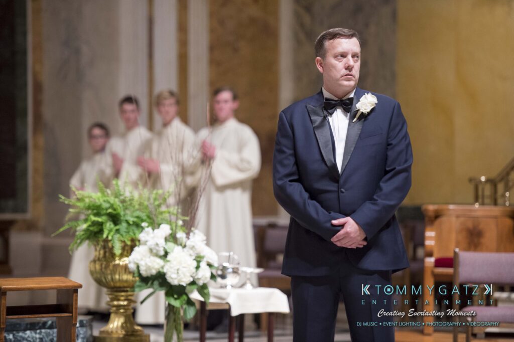 © alexandermorozovcom All Rights Reserved inquire by text +1-443-858-8743 - TOMMY GATZ ENTERTAINMENT A man in a formal suit stands in a church setting, looking slightly to the side. People in white robes are blurred in the background near a floral arrangement.