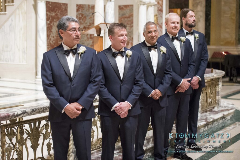 Elizabeth-Tom-101417-CEREMONY-Low-Res-WM-204 - TOMMY GATZ ENTERTAINMENT Five men in matching tuxedos stand in a line indoors, smiling, with floral boutonnieres. Marble columns and ornate railing are in the background.