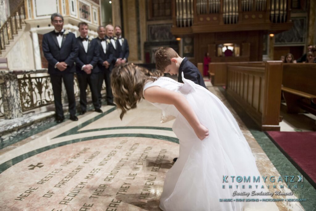Elizabeth-Tom-101417-CEREMONY-Low-Res-WM-213 - TOMMY GATZ ENTERTAINMENT Two children in formal attire bow in front of a church altar during a ceremony, with groomsmen standing in the background.