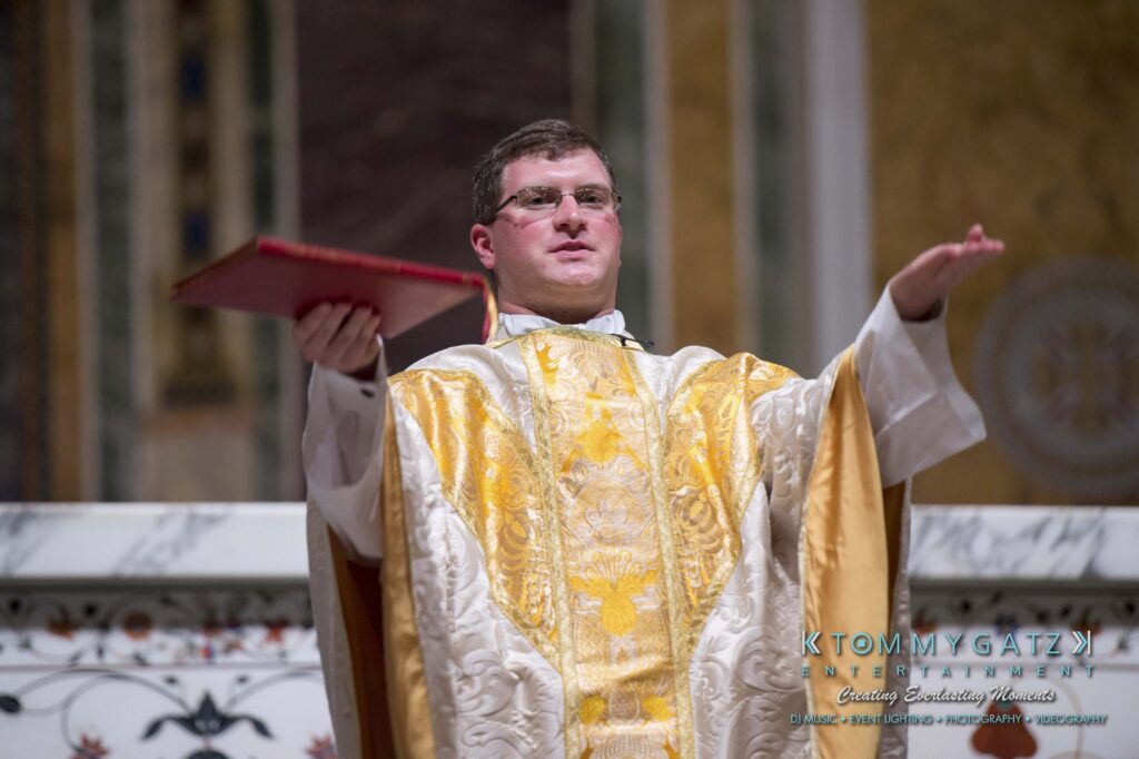 © alexandermorozovcom All Rights Reserved inquire by text +1-443-858-8743 - TOMMY GATZ ENTERTAINMENT A priest in yellow and white vestments holds an open book while speaking, standing in a church setting with ornate decor.