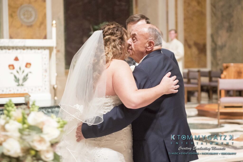 Elizabeth-Tom-101417-CEREMONY-Low-Res-WM-228 - TOMMY GATZ ENTERTAINMENT A bride in a white gown and veil hugs an older man in a suit inside a church. A religious altar is visible in the background.