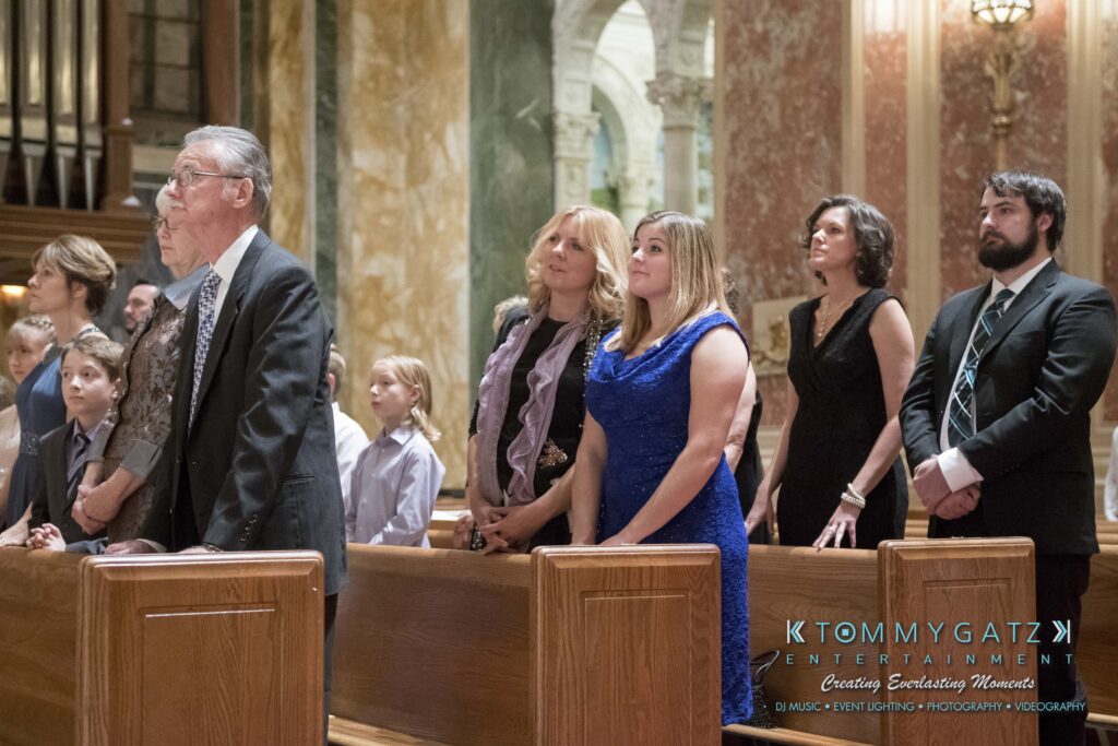 Elizabeth-Tom-101417-CEREMONY-Low-Res-WM-236 - TOMMY GATZ ENTERTAINMENT A group of people stand in a church pew during a service. They are dressed formally, with men in suits and women in dresses.