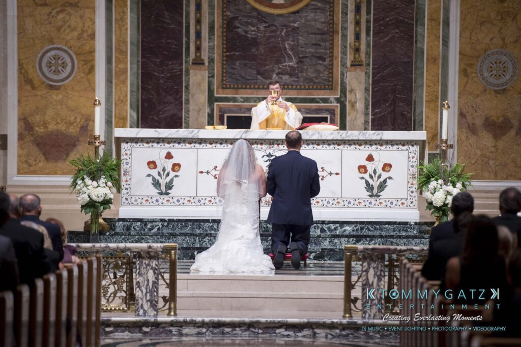 © alexandermorozovcom All Rights Reserved inquire by text +1-443-858-8743 - TOMMY GATZ ENTERTAINMENT A couple kneels before a priest at an ornate altar during a wedding ceremony, with guests seated in the background.