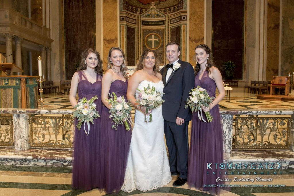 Elizabeth-Tom-101417-FORMALS-Low-Res-WM-20 - TOMMY GATZ ENTERTAINMENT A wedding couple poses with four bridesmaids in purple dresses, each holding a bouquet, inside an ornate church setting.