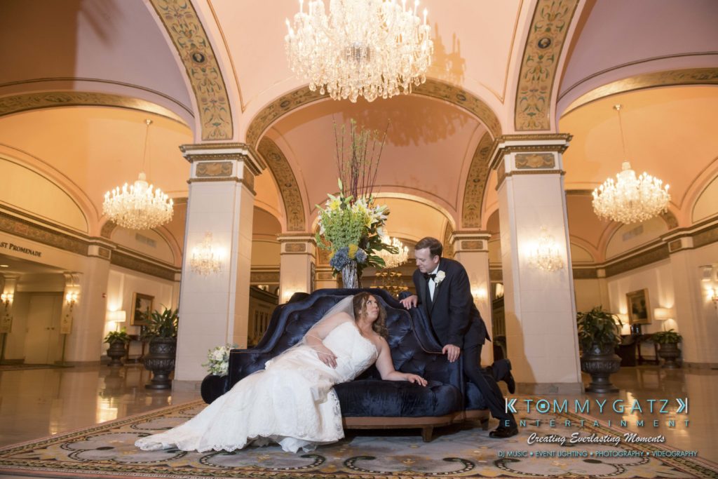 Elizabeth-Tom-101417-FORMALS-Low-Res-WM-48-1024x683 - TOMMY GATZ ENTERTAINMENT Bride and groom in formal attire pose together on a blue sofa in an ornate ballroom with chandeliers and floral arrangements.
