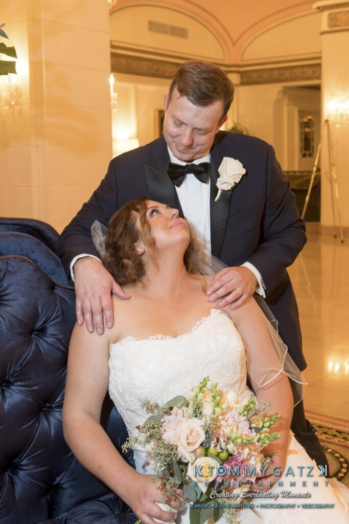 Elizabeth-Tom-101417-FORMALS-Low-Res-WM-58 - TOMMY GATZ ENTERTAINMENT A bride and groom in wedding attire pose indoors. The bride holds a bouquet and sits, looking up at the groom, who stands behind with a hand on her shoulder.