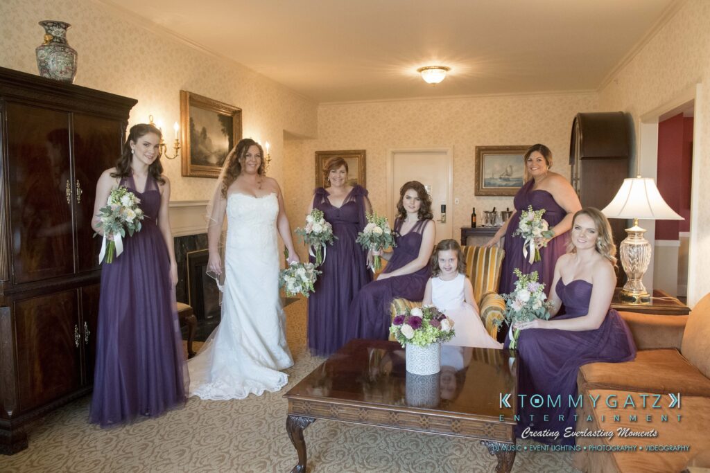 Elizabeth-Tom-101417-PREP-Low-Res-WM-10 - TOMMY GATZ ENTERTAINMENT Bride with five bridesmaids in purple dresses and a young girl in white, posing in a warmly furnished room. The bridesmaids hold flower bouquets.