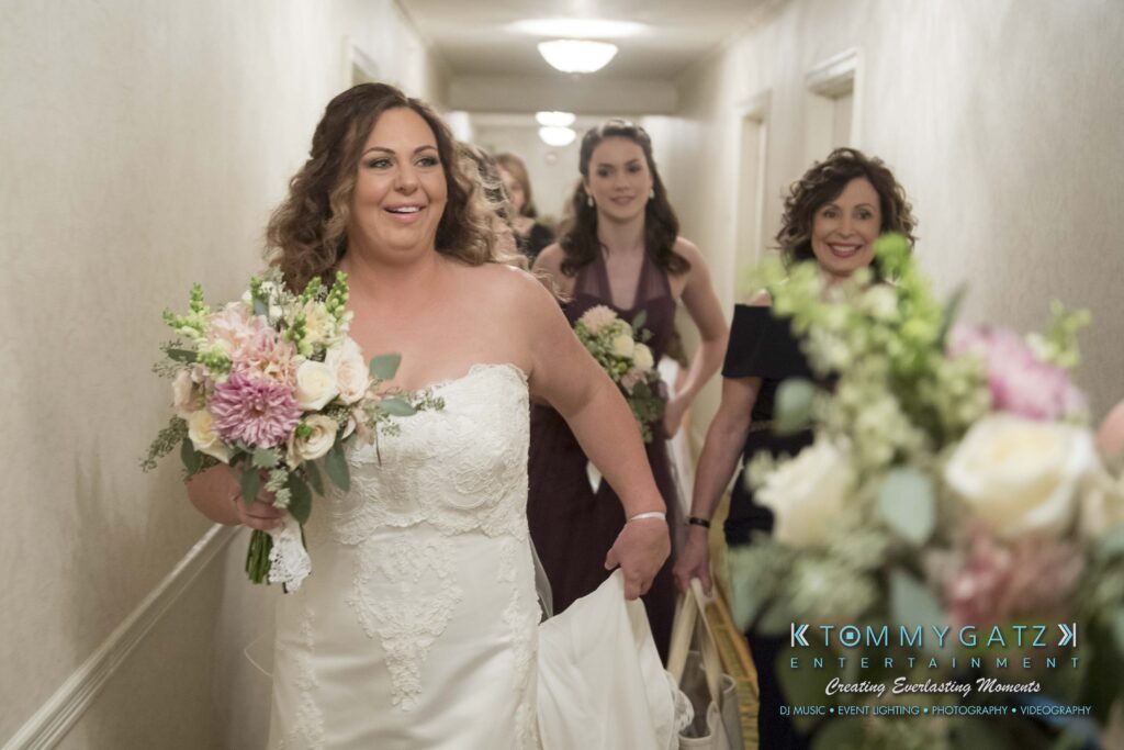 Elizabeth-Tom-101417-PREP-Low-Res-WM-36 - TOMMY GATZ ENTERTAINMENT A bride in a white dress walks with a bouquet, accompanied by smiling women in an indoor corridor.