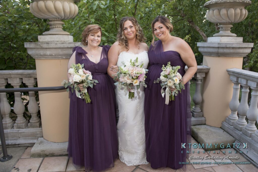 Elizabeth-Tom-101417-PREP-Low-Res-WM-59 - TOMMY GATZ ENTERTAINMENT A bride stands between two bridesmaids on a terrace. All are holding bouquets, and the bridesmaids wear matching purple dresses.