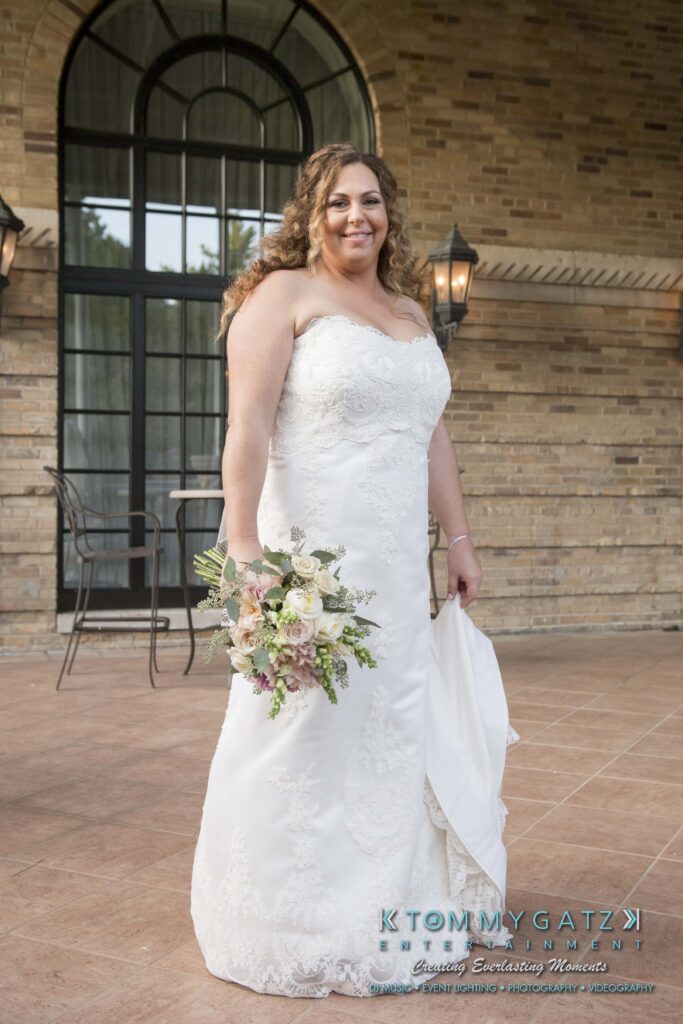 © alexandermorozovcom All Rights Reserved inquire by text +1-443-858-8743 - TOMMY GATZ ENTERTAINMENT A woman in a white wedding dress holds a bouquet of flowers standing outside a brick building.
