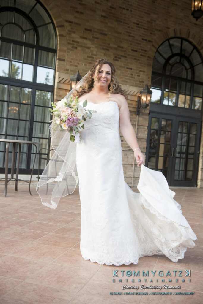 Elizabeth-Tom-101417-PREP-Low-Res-WM-71 - TOMMY GATZ ENTERTAINMENT A bride in a white dress and veil holds a bouquet while standing on a patio. She is smiling and holding her dress with one hand. The background features arched windows and brickwork.