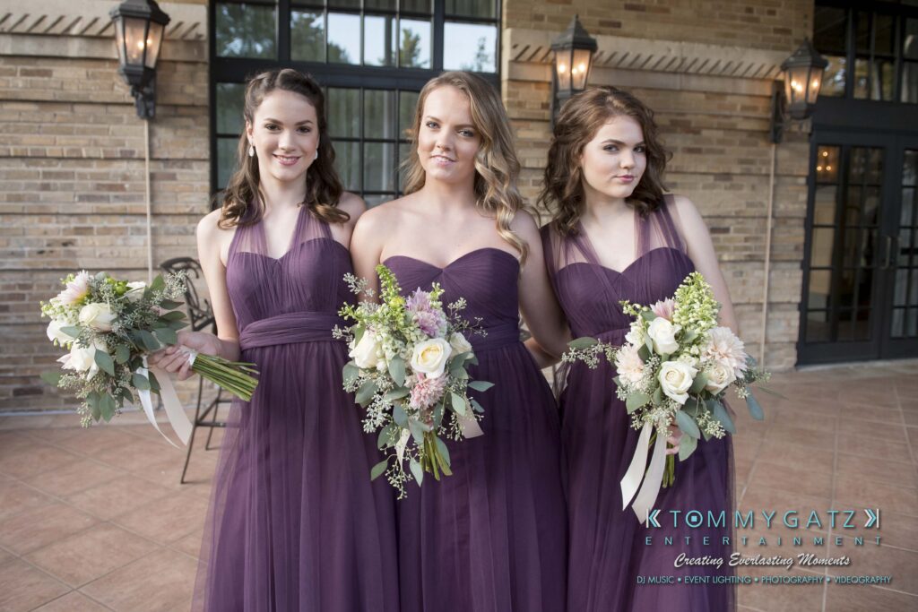 Elizabeth-Tom-101417-PREP-Low-Res-WM-76 - TOMMY GATZ ENTERTAINMENT Three women in matching purple dresses hold bouquets while standing in front of a brick wall.