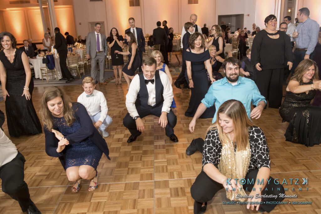 Elizabeth-Tom-101417-RECEPTION-Low-Res-WM-204 - TOMMY GATZ ENTERTAINMENT People on a dance floor participating in a group dance. Some are kneeling with hands out, while others are standing. Tables and other guests are visible in the background.