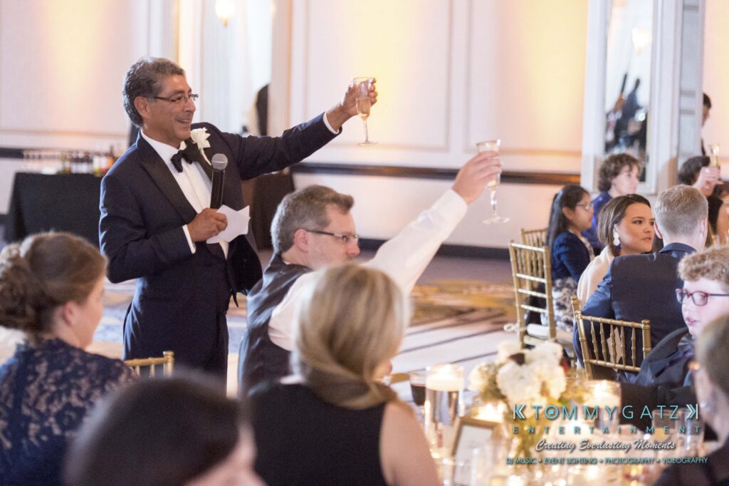 © alexandermorozovcom All Rights Reserved inquire by text +1-443-858-8743 - TOMMY GATZ ENTERTAINMENT A man in a tuxedo gives a toast with a raised glass at a formal event. Guests seated at tables listen and raise their glasses in response.