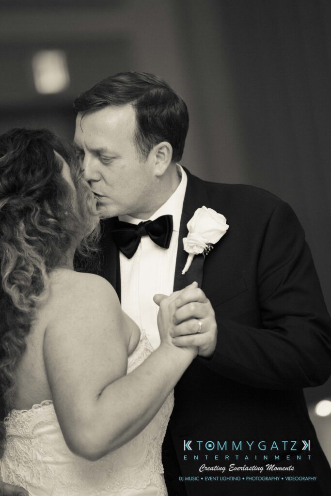 Elizabeth-Tom-101417-RECEPTION-Low-Res-WM-31 - TOMMY GATZ ENTERTAINMENT A couple dressed in wedding attire share a kiss on the dance floor. The groom wears a tuxedo with a bow tie and boutonnière, while the bride has curly hair and a strapless gown.