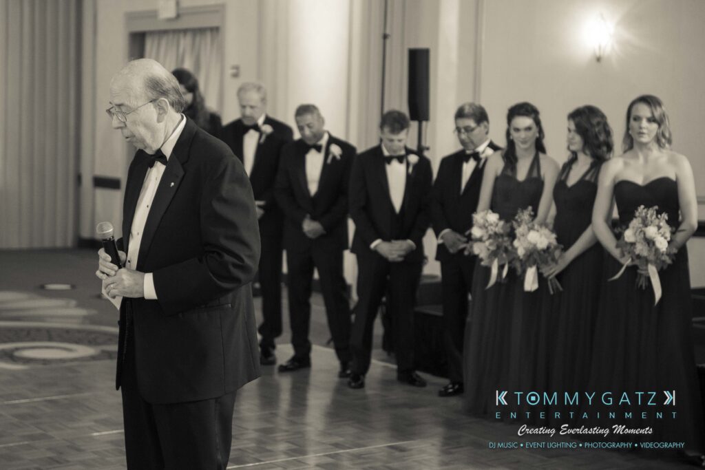 Elizabeth-Tom-101417-RECEPTION-Low-Res-WM-405 - TOMMY GATZ ENTERTAINMENT An elderly man in a suit speaks into a microphone. A group of people in formal wear stands in the background, some holding bouquets.