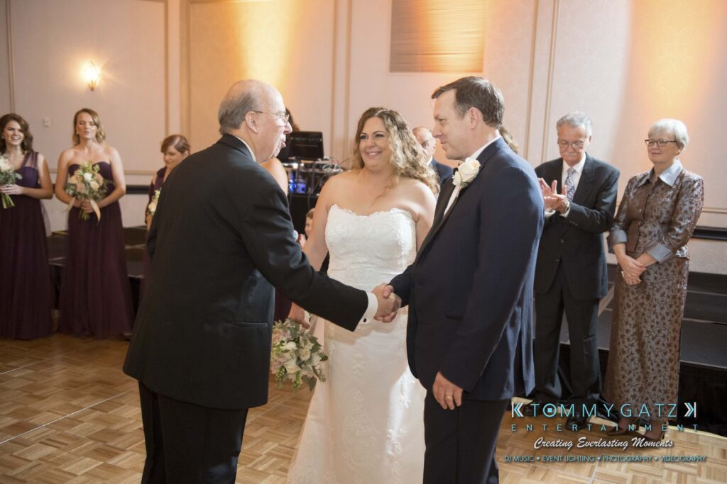 © alexandermorozovcom All Rights Reserved inquire by text +1-443-858-8743 - TOMMY GATZ ENTERTAINMENT A bride in a white dress smiles as a man in a black suit shakes hands with a groom in a blue suit during a wedding ceremony, with bridesmaids and guests in the background.