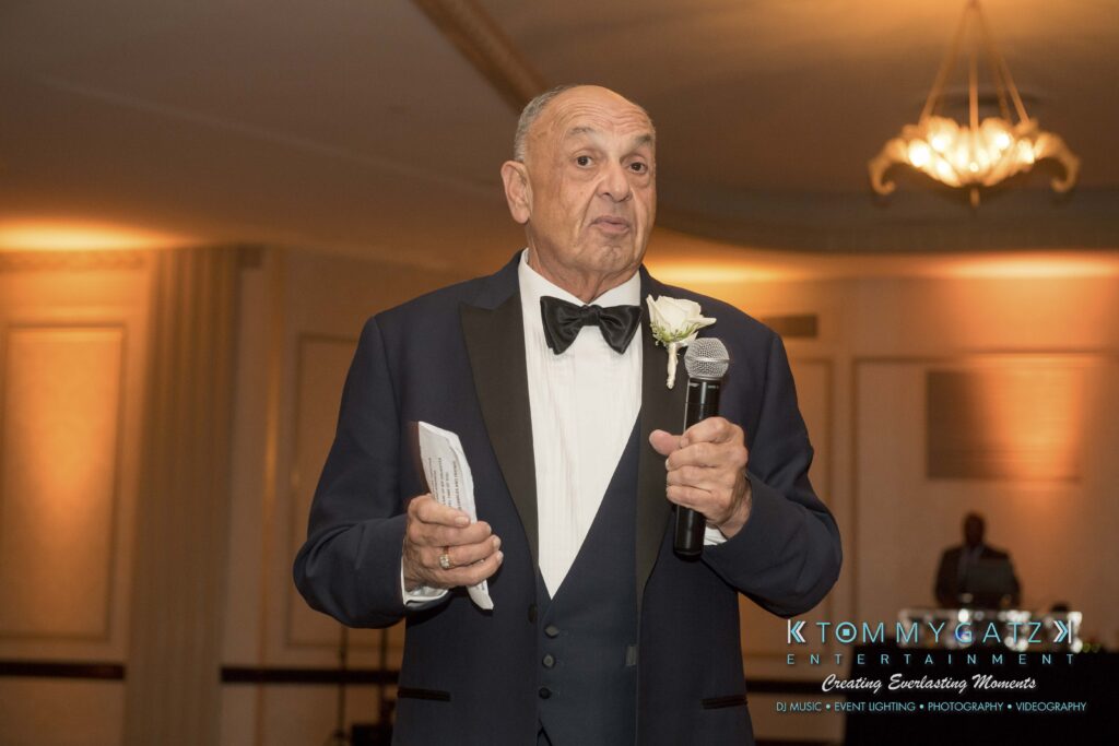 Elizabeth-Tom-101417-RECEPTION-Low-Res-WM-5 - TOMMY GATZ ENTERTAINMENT An elderly man in a tuxedo is holding a microphone and speaking at an indoor event with chandelier lighting.