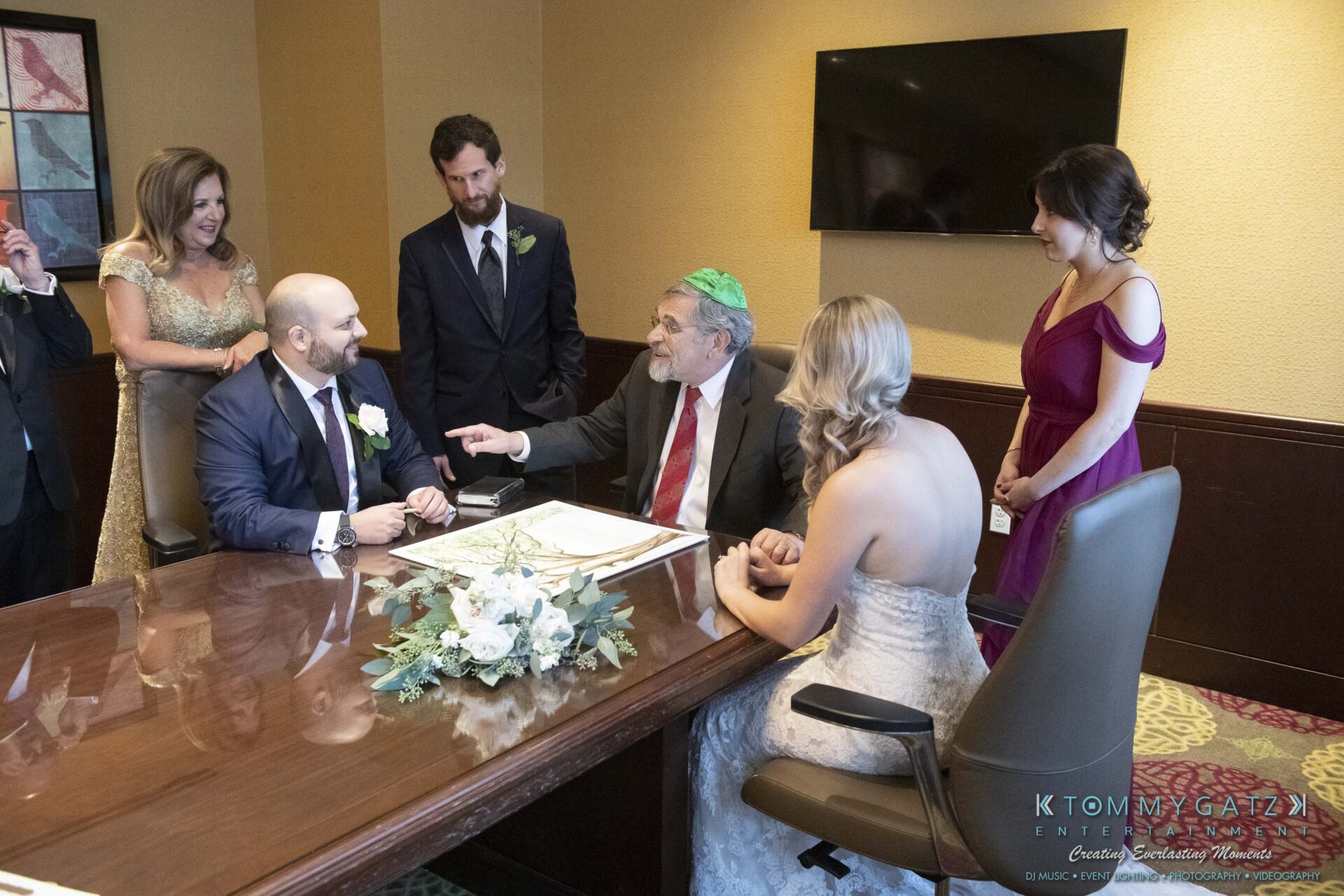 A group of people in formal attire sit and stand around a wooden table with documents. A man wearing a green kippah gestures while others listen, including a person in a white wedding dress.