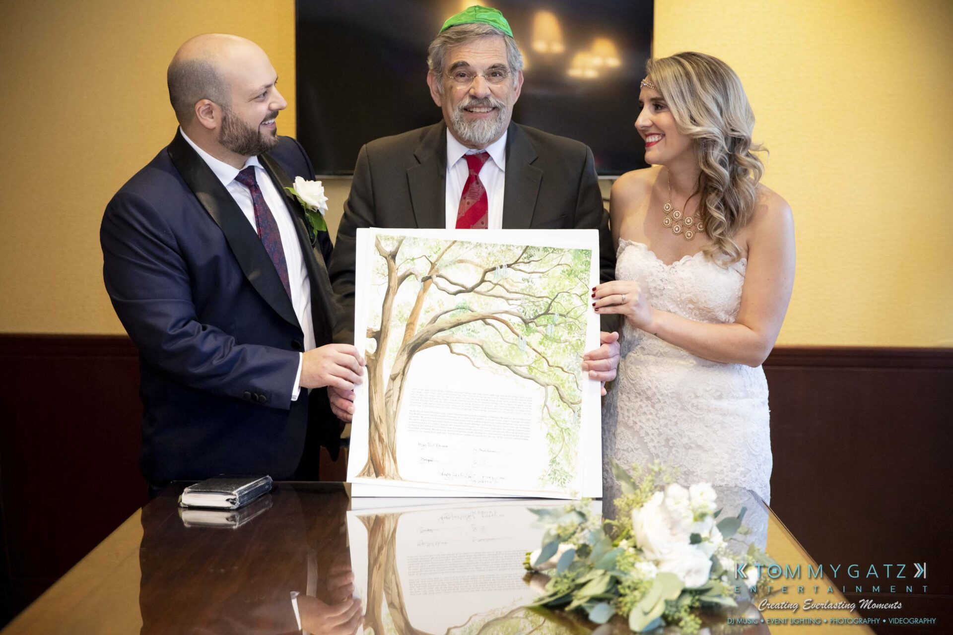Three people stand together indoors. The middle person holds a framed picture of a tree. A bouquet rests on the table in front of them.