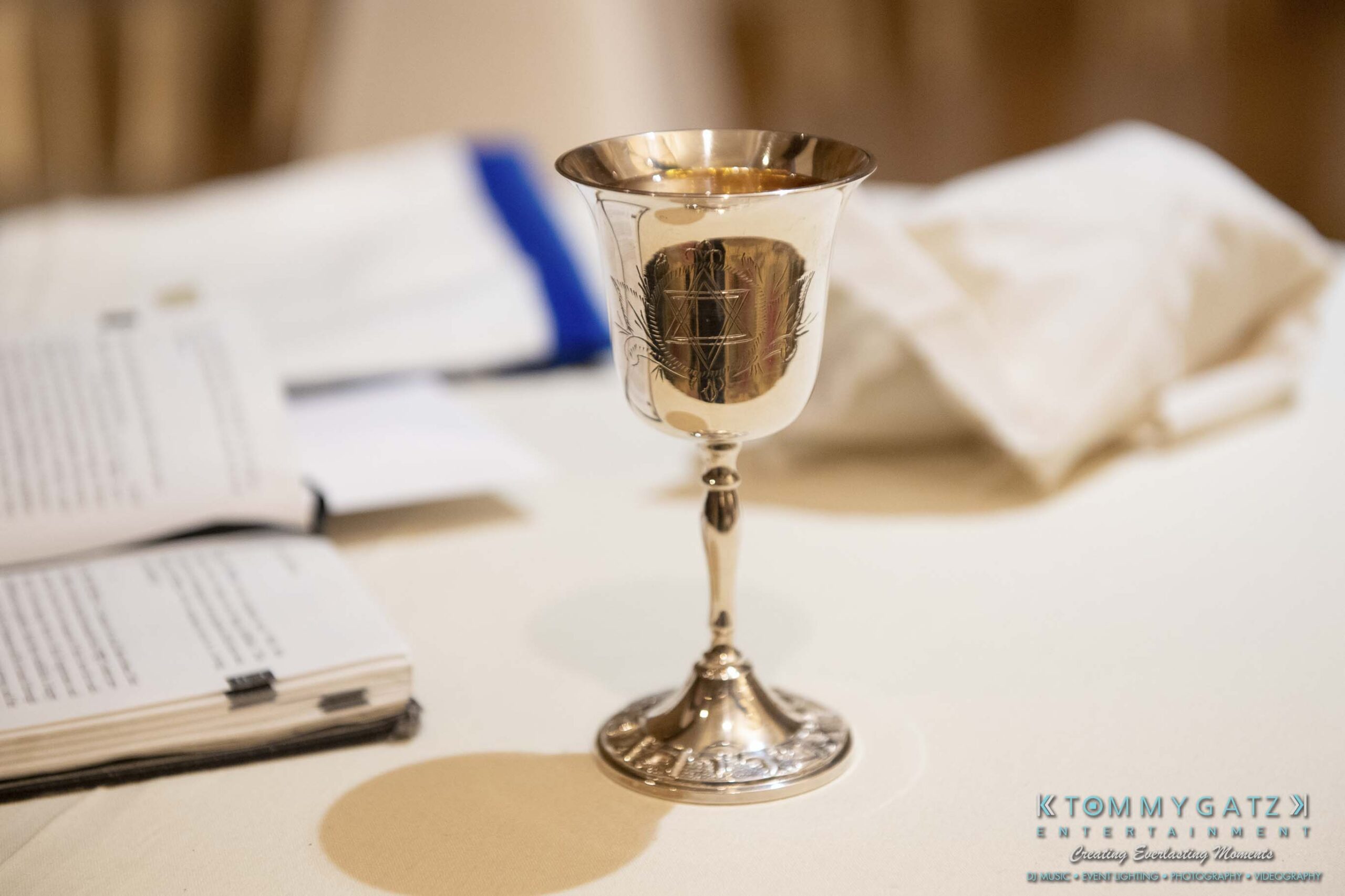 A silver goblet with ornate etchings is placed on a table beside an open book and folded white cloth.