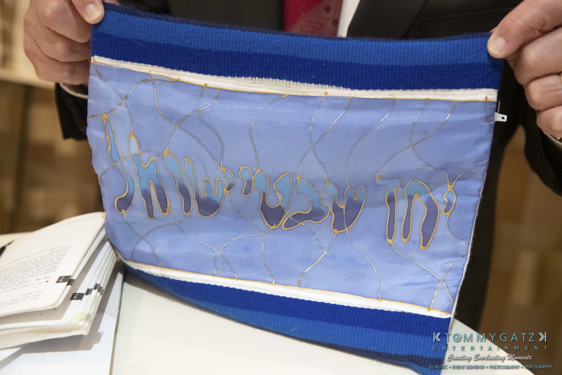 Person holding a blue fabric piece with silver and gold embroidered Hebrew text.