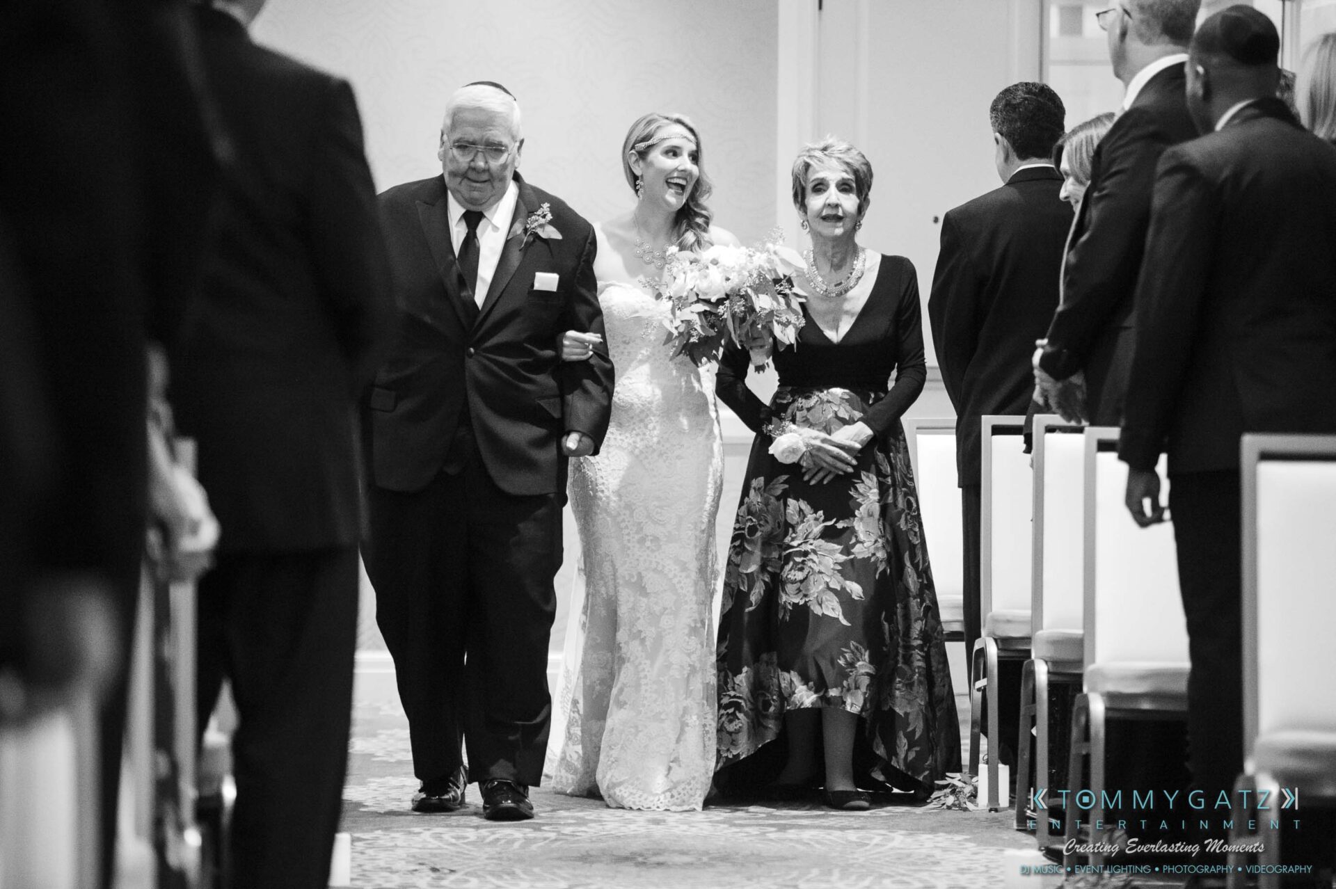 A bride in a wedding dress, arm-in-arm with two elderly people, walks down the aisle in a ceremony.