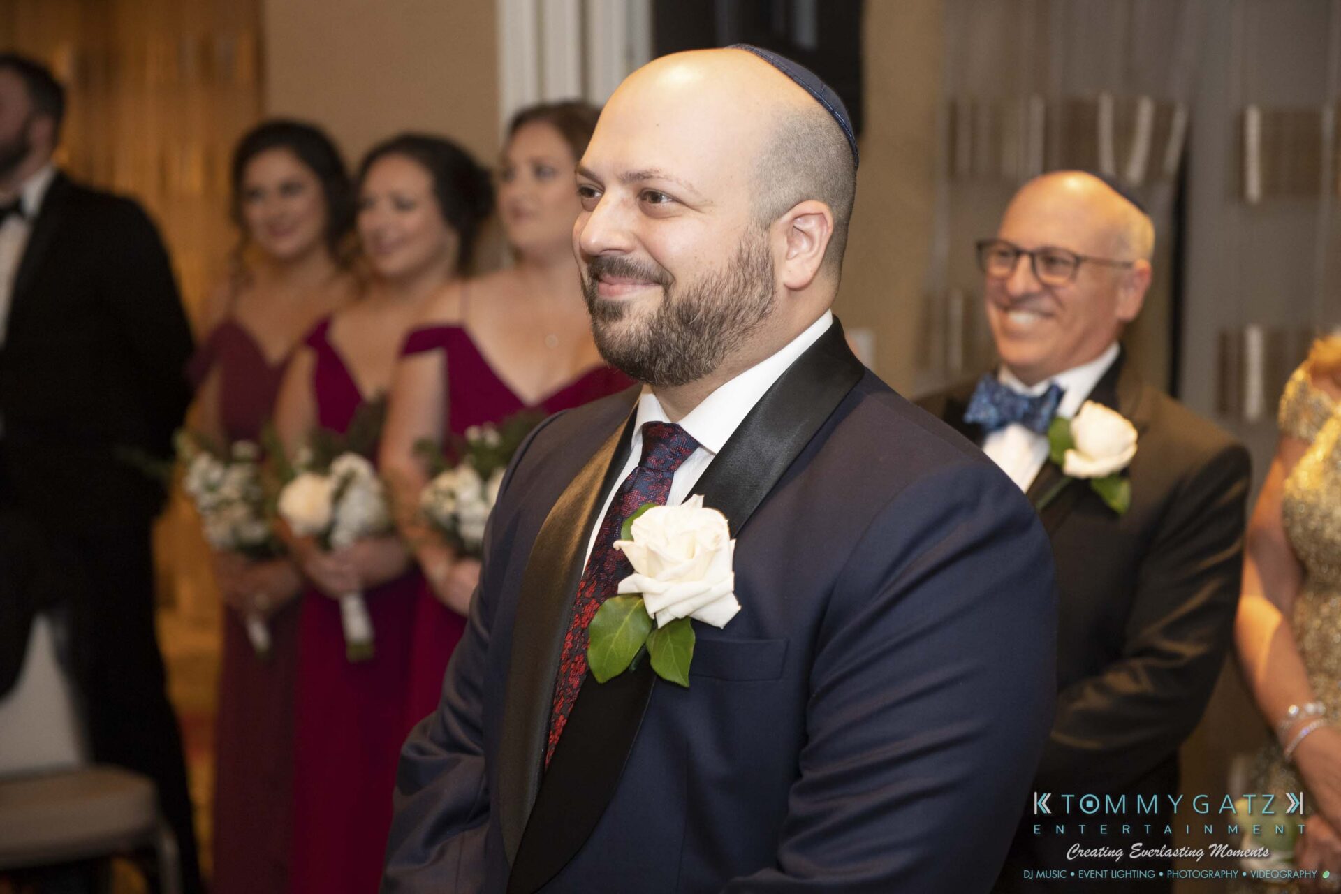 A groom in a blue suit stands in a room with a group of people in formal attire, including bridesmaids in maroon dresses and a man in glasses with a bow tie.