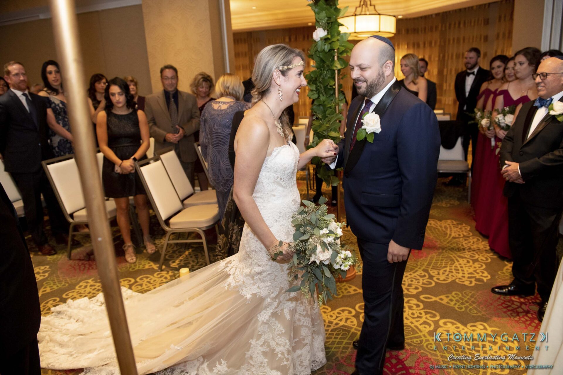 A bride and groom hold hands during their wedding ceremony, surrounded by guests in formal attire.