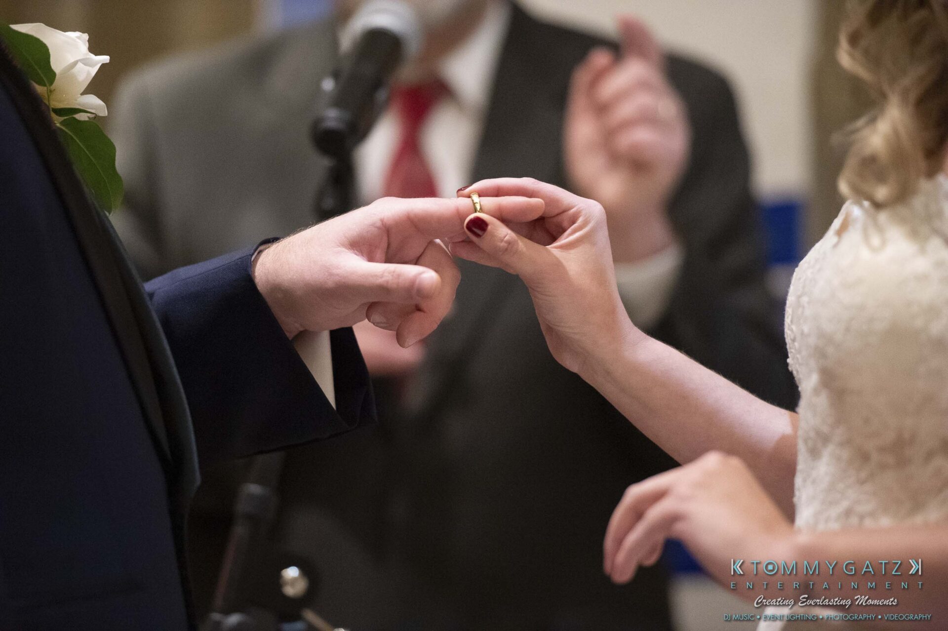 Close-up of a bride placing a ring on the groom's finger during a wedding ceremony, with an officiant in the background.