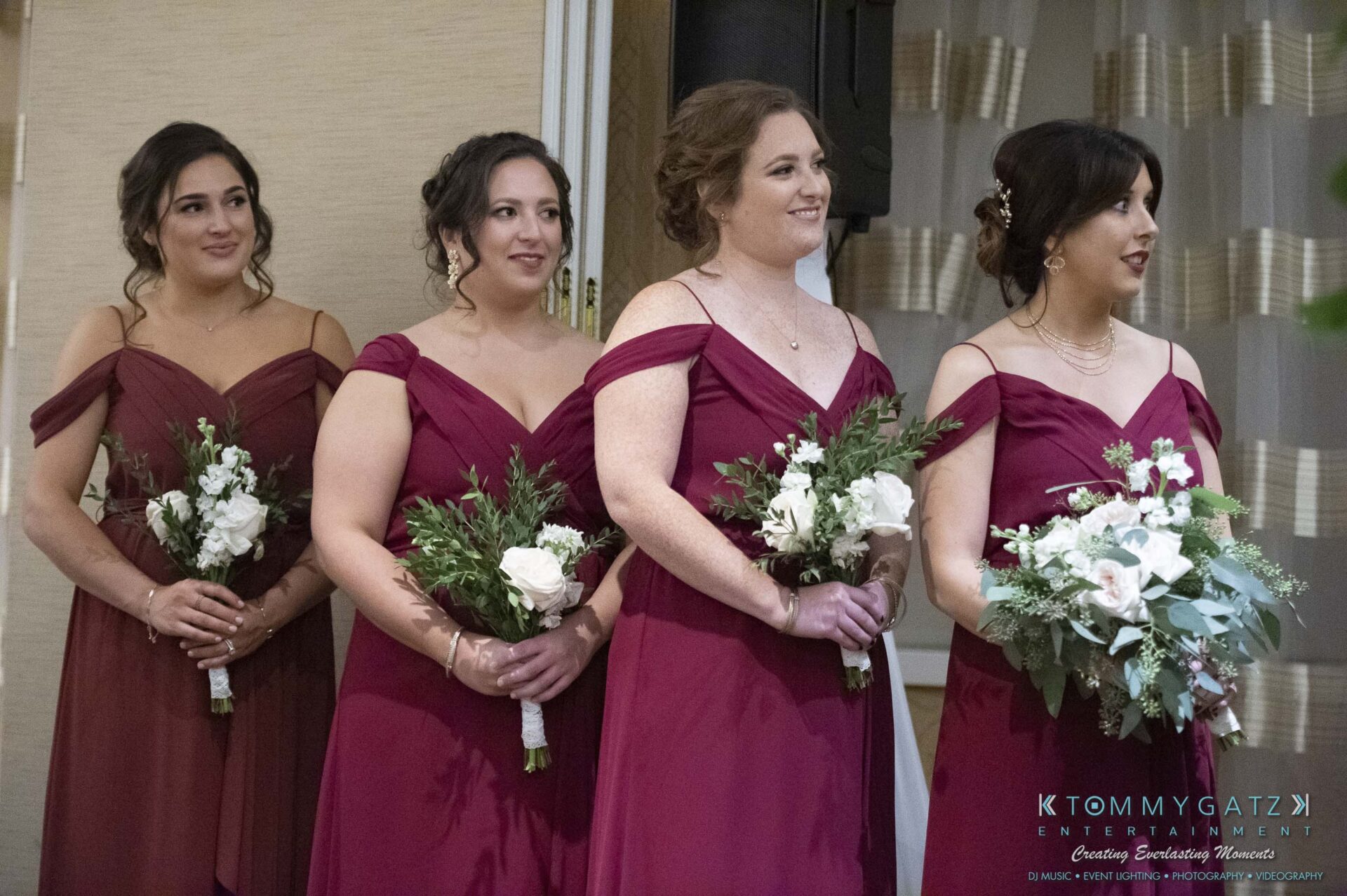 Four women in matching burgundy dresses stand in a row holding bouquets, smiling at an indoor event.