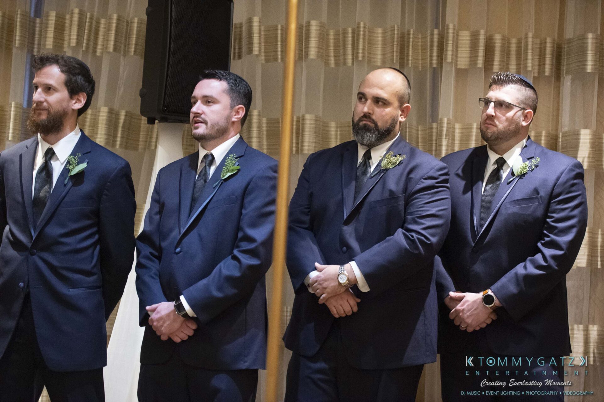 Four men in suits stand side by side indoors, each wearing a boutonniere.