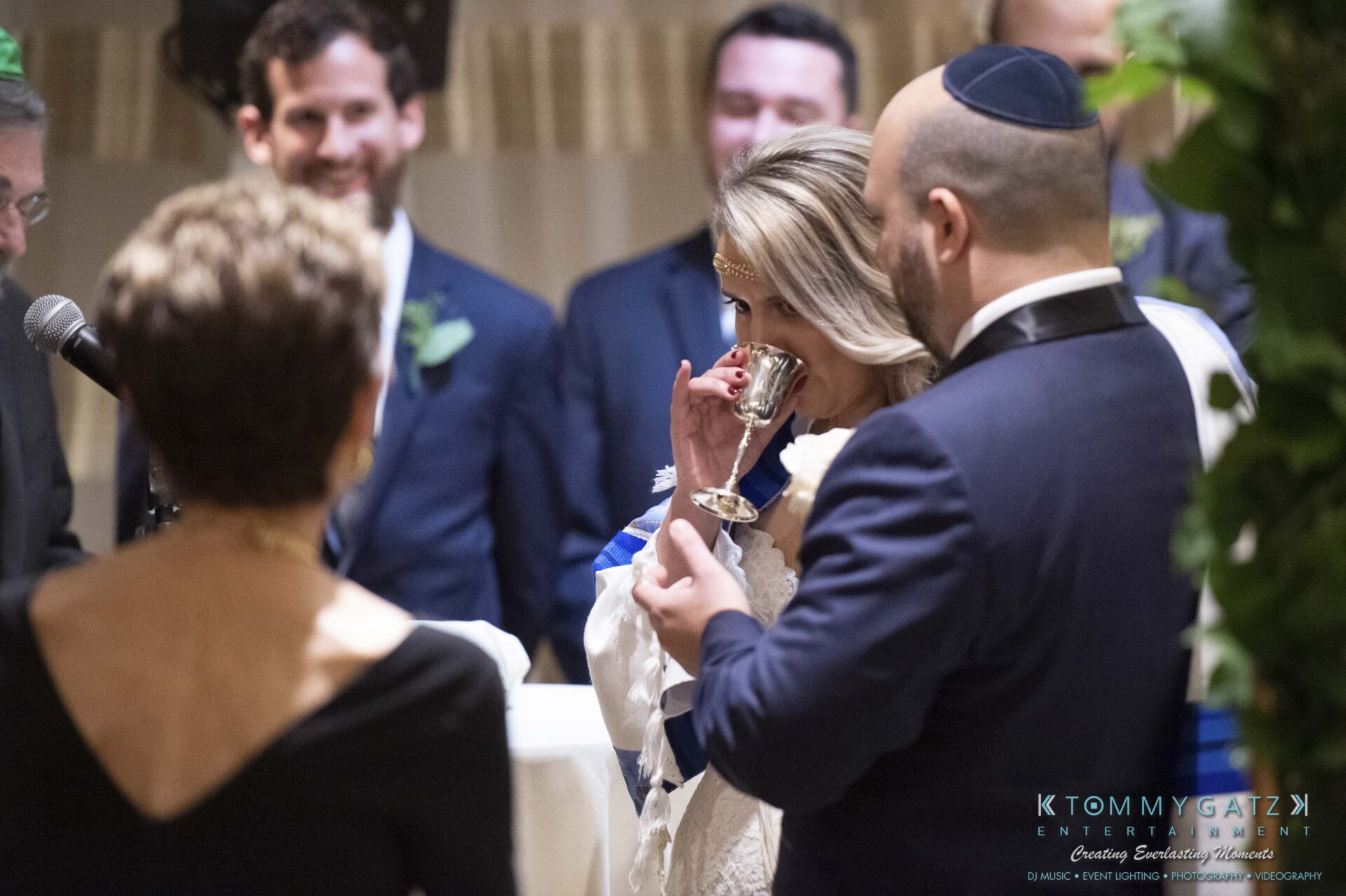 A couple participates in a wedding ceremony surrounded by people, while the bride drinks from a cup.
