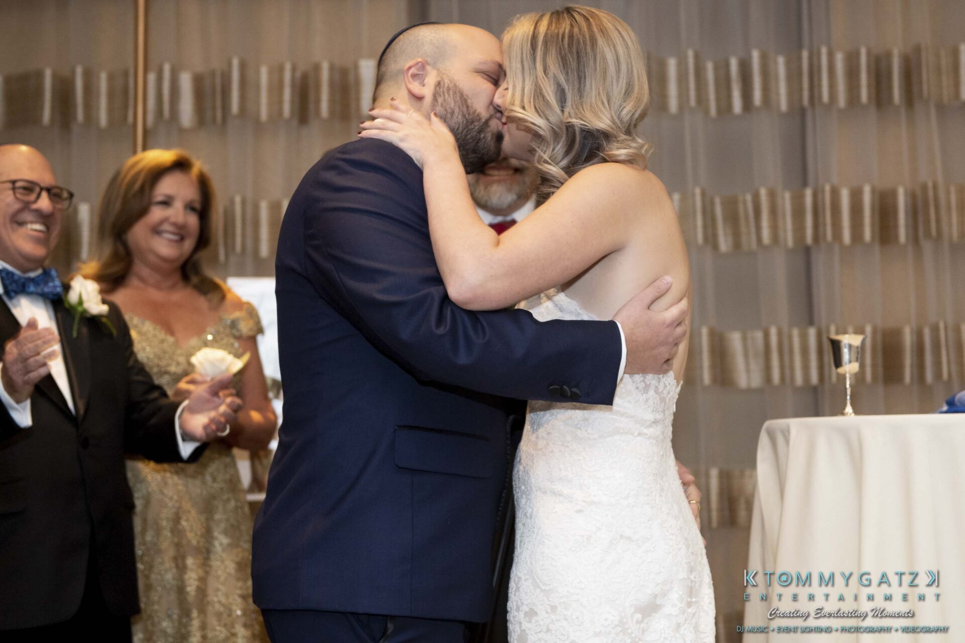 A couple kisses at their wedding ceremony while guests smile and applaud in the background.