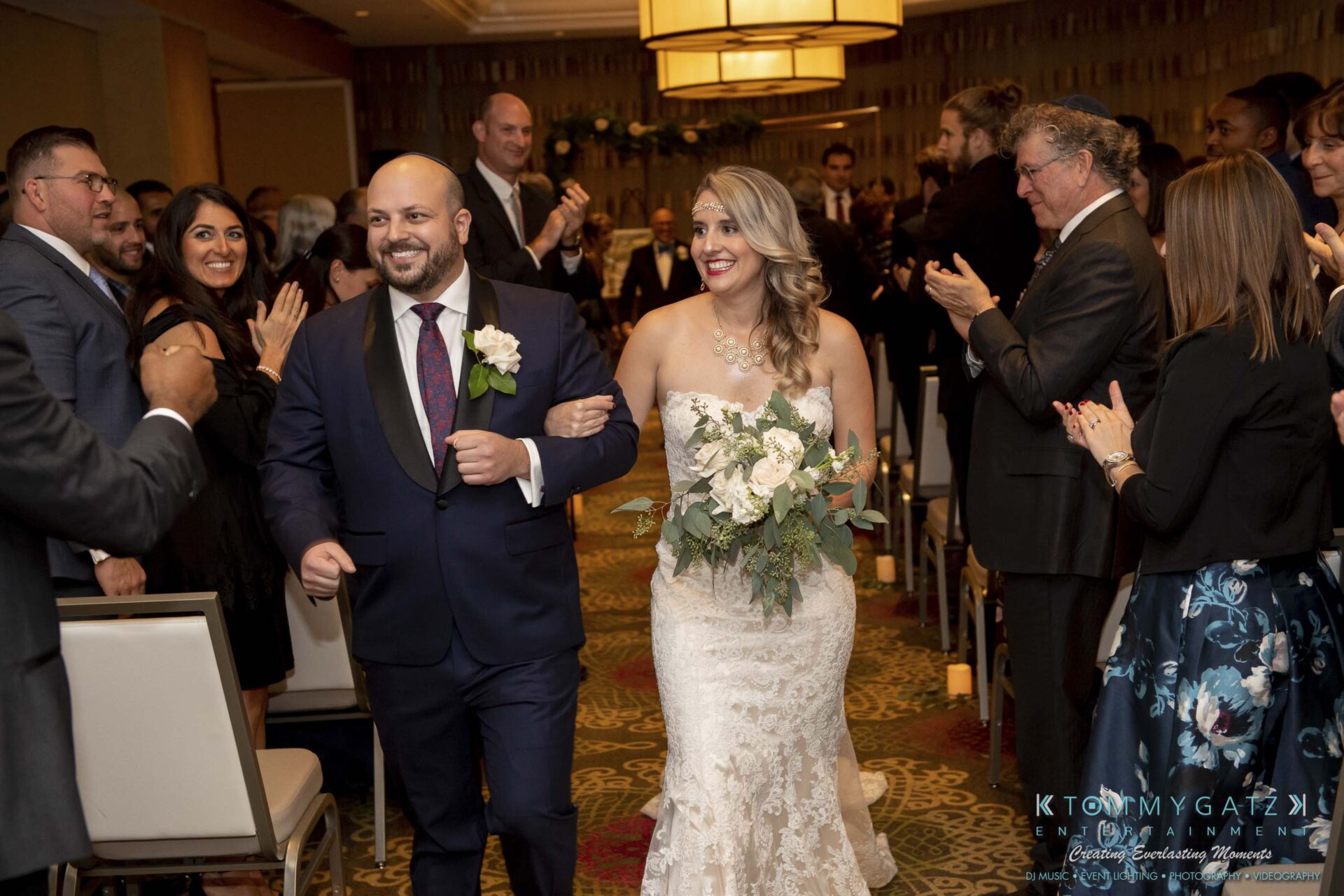 A smiling couple walks down the aisle, with the groom in a suit and the bride in a lace gown. Guests are clapping and standing in the background.